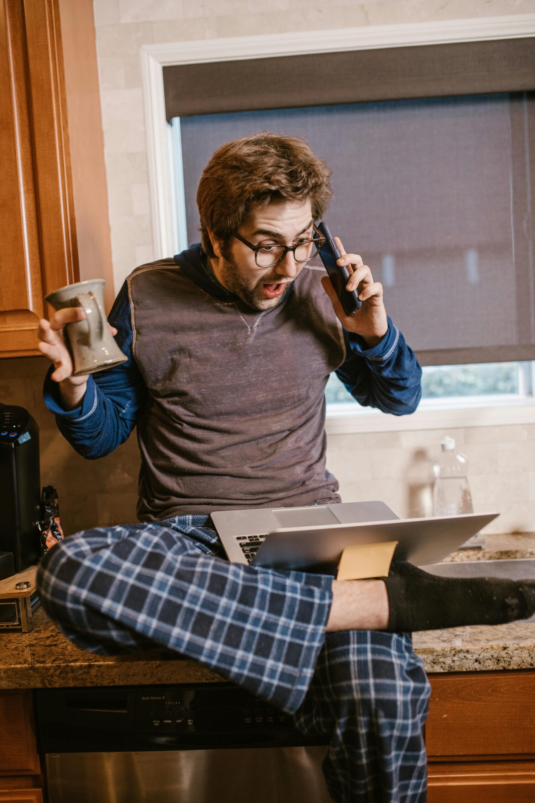 Man in pajamas shocked while on the phone and using a laptop, sitting on kitchen counter.